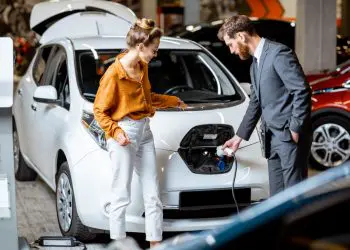 Sales manager showing how to charge electric car to a young client, plugging wire into the car socket at the car dealership