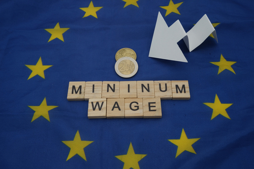 Some coins, next to a white arrow and a wooden inscription, minimum wage, above a flag of the European Union.