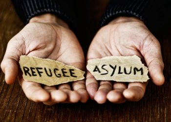 closeup of the hands of a young man with two pieces of paper with the words refugee and asylum written in each one, with a dramatic effect