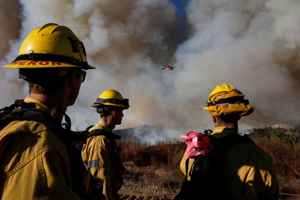 Firefighters watch as a helicopter flies through smoke